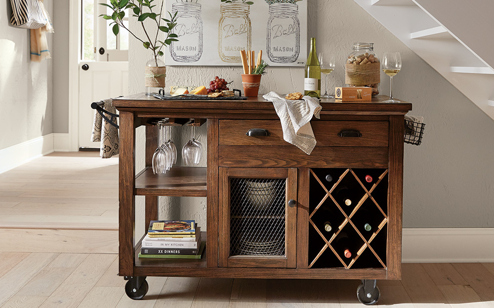 A brown wooden bar cart stores wine, glassware and dinnerware while doubling as a serving cart for a cheese board.