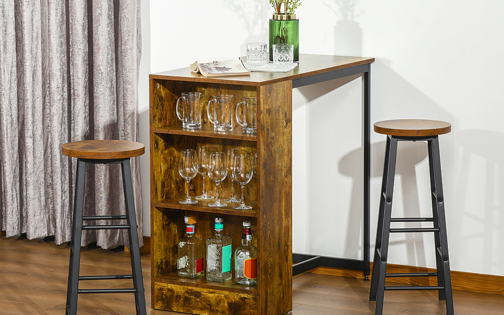 A wooden bar table with shelves holds glassware and spirits and stands between two wood and metal stools.