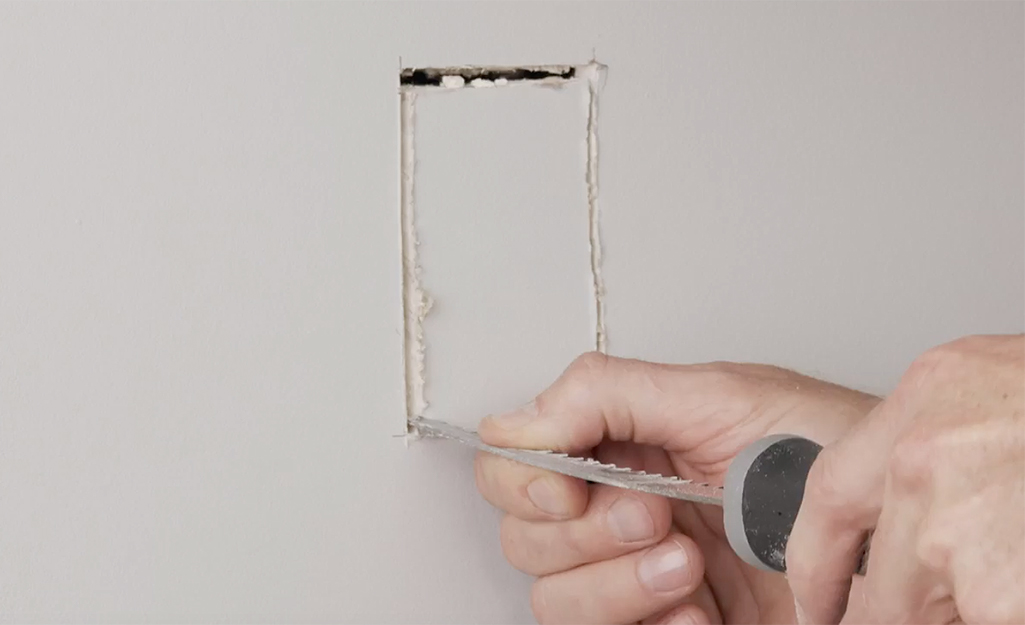A person cutting a rectangle into drywall.