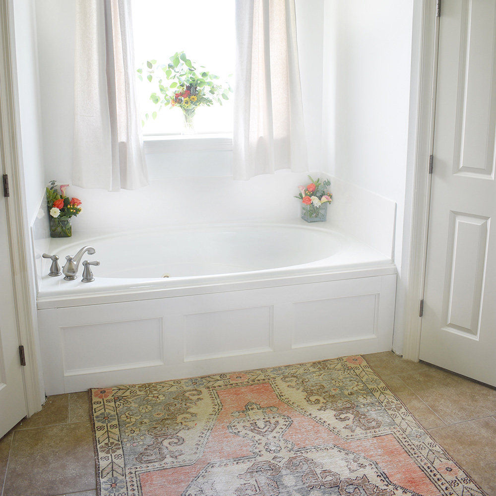 A bathroom with patterned rug in front of a white bathtub.