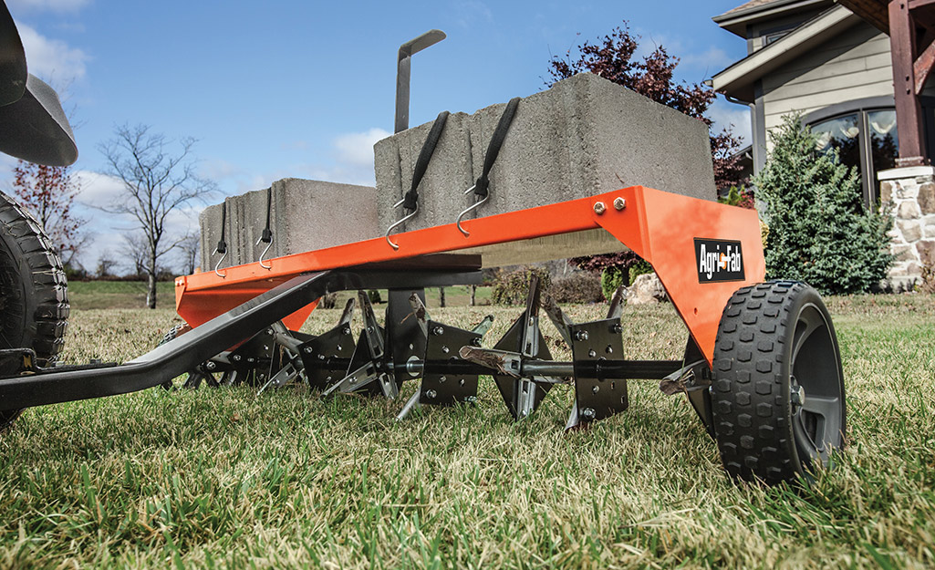 A lawn aerator weighed down by cement blocks being pulled over an unhealthy looking lawn.