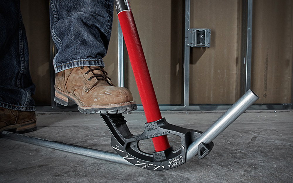 A person uses his boot to steady a conduit bender as it bends a pipe.