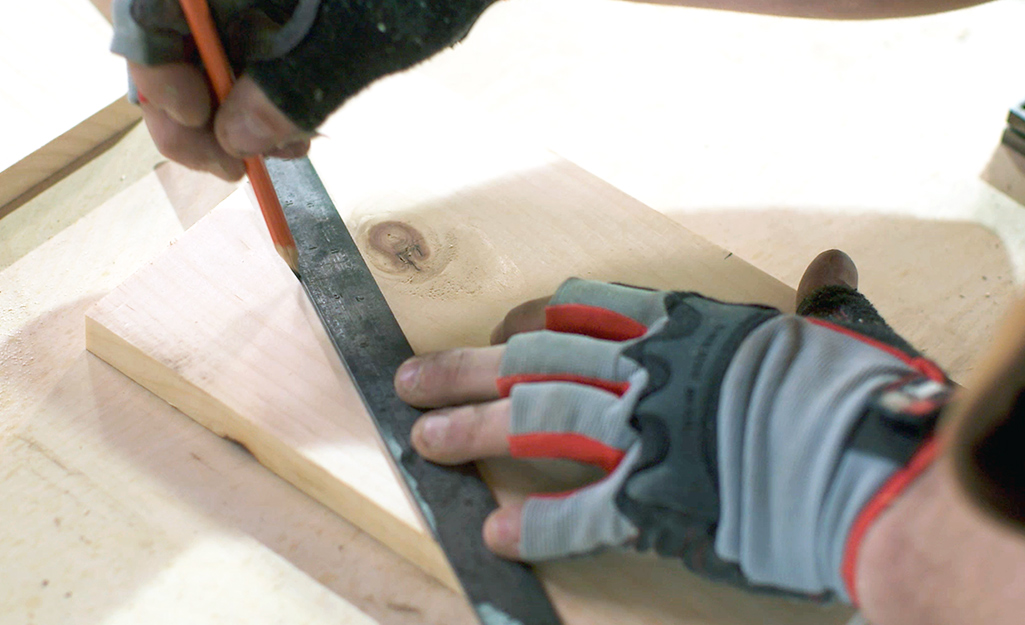 A person uses a pencil and a ruler to mark a cut line on a piece of wood needed to build the birdhouse.