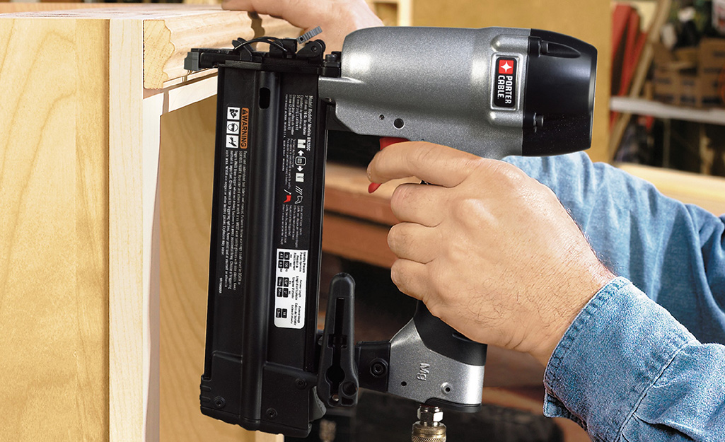 Someone using a finish nailer to fasten moulding along the top of a bookshelf.