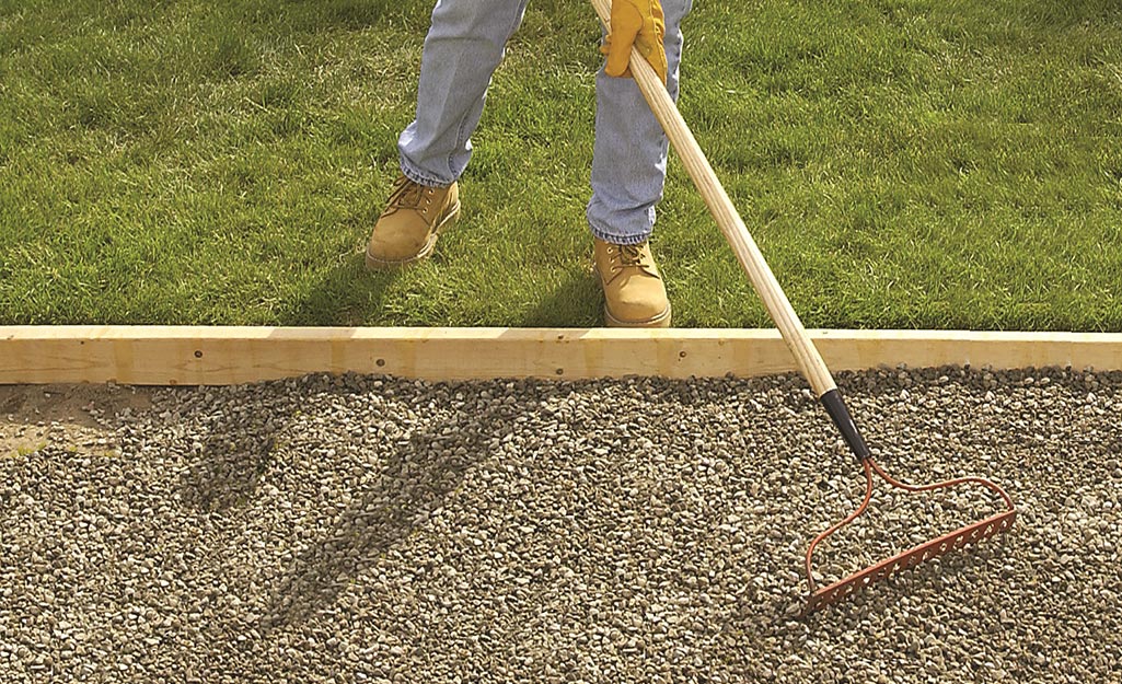 A person uses a metal rake to flatten an area of gravel ground cover.