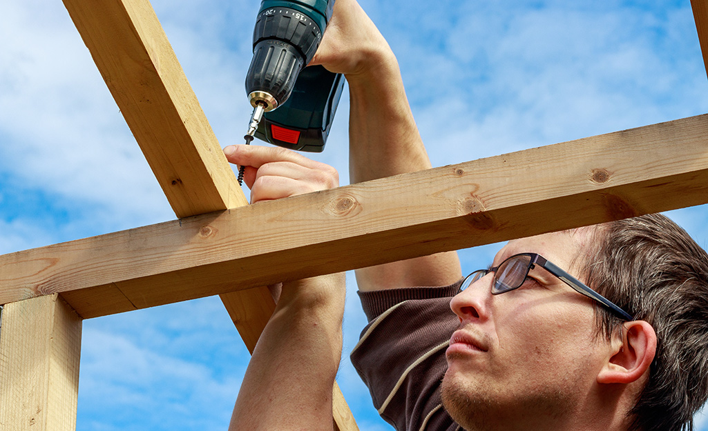 A person uses a power drill to attach a rafter to a beam.