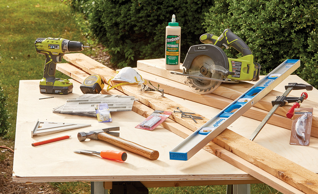 A worktable displays the tools and materials needed to make a cold frame, including a hammer, a level, a drill and a circular saw.