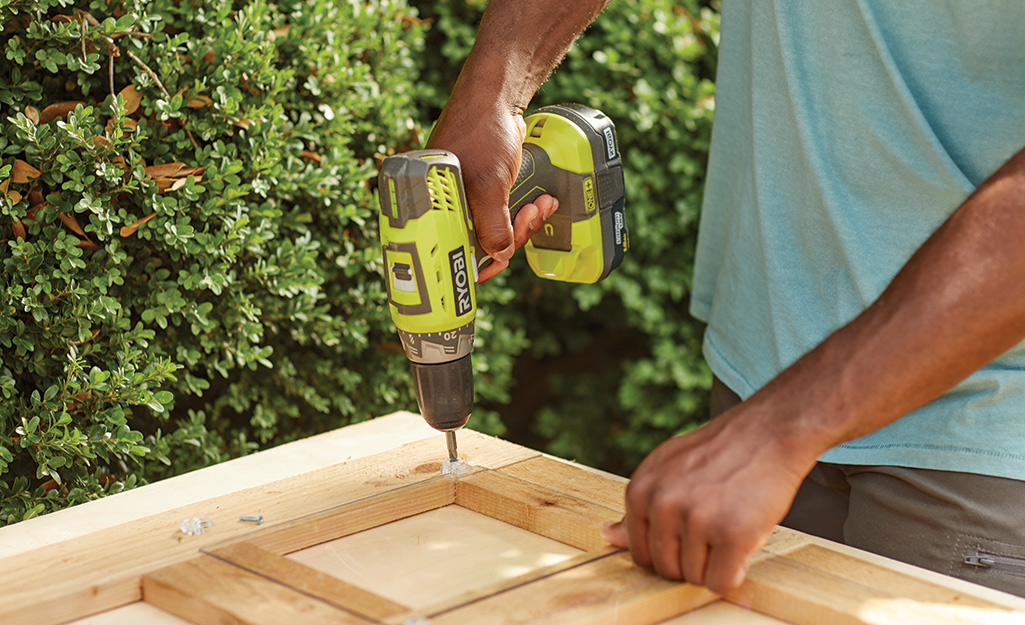 A person uses a drill to attach screws to the corner of a cold frame. 