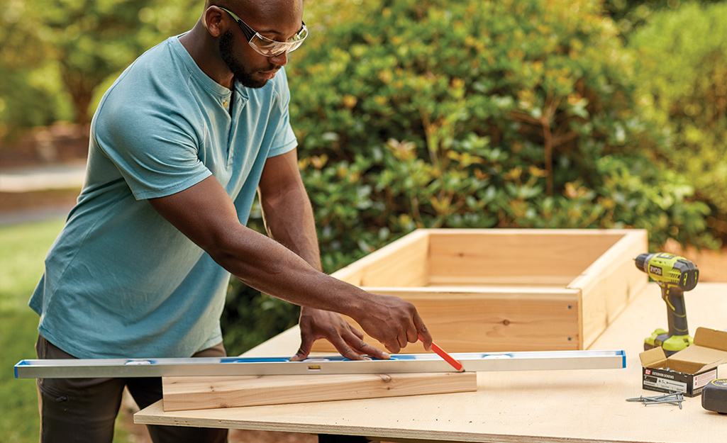 A man uses a straight edge and a pencil to draw a cut line on a board for a wedge of a cold frame.
