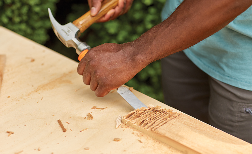 A person uses a hammer and a chisel to chip wood off a board that will be used to make the top of a cold frame.