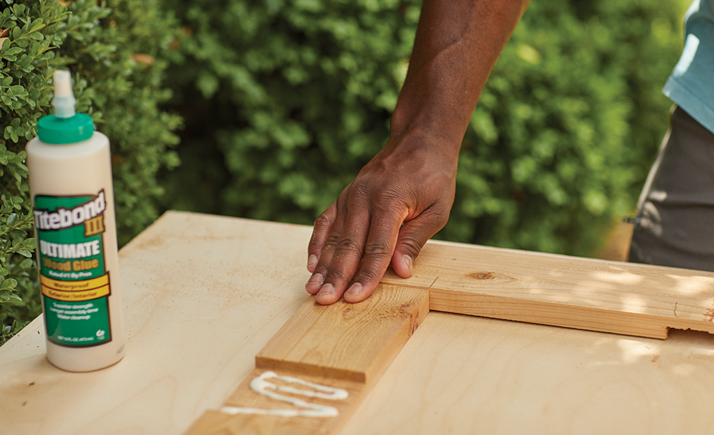 A person holds down a small piece of wood that is being glued to a larger piece on the top part of a cold frame.