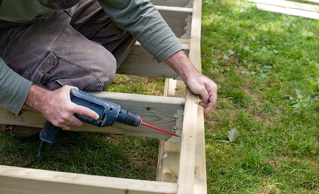 Man using a power drill to attach a deck frame's inner joist. Man using a power drill to attach a deck frame's inner joist.