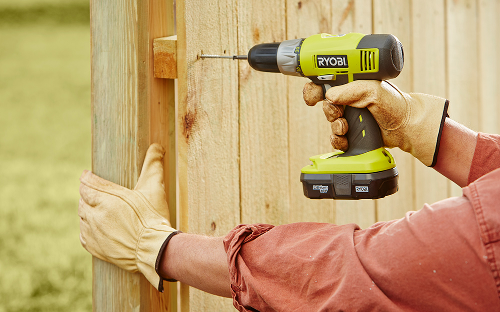 A person uses a power drill to secure a rail to a fence. 