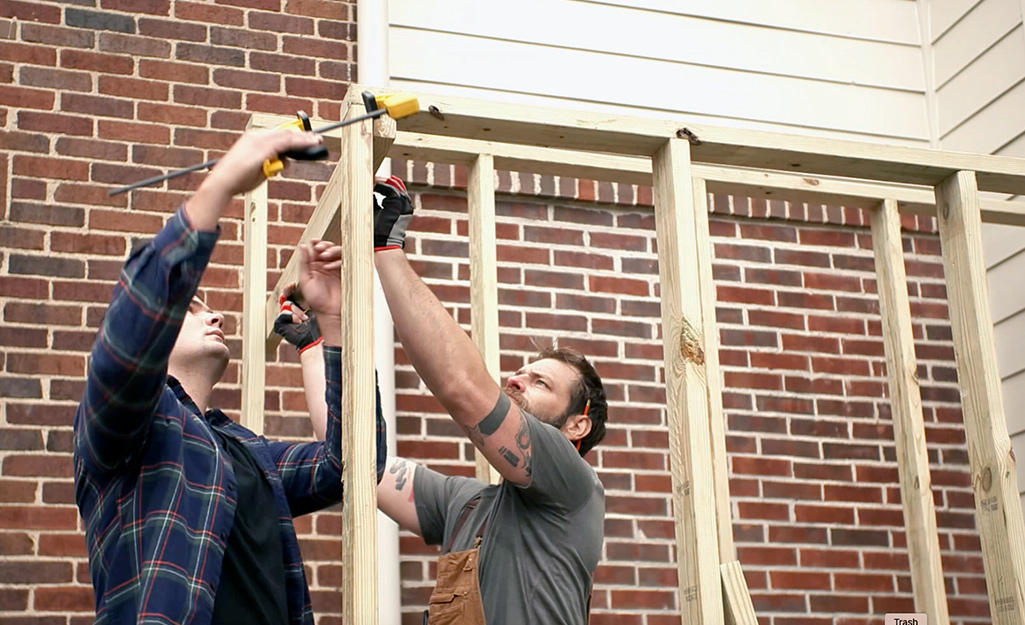 Two men work on building the frame of a wooden greenhouse.