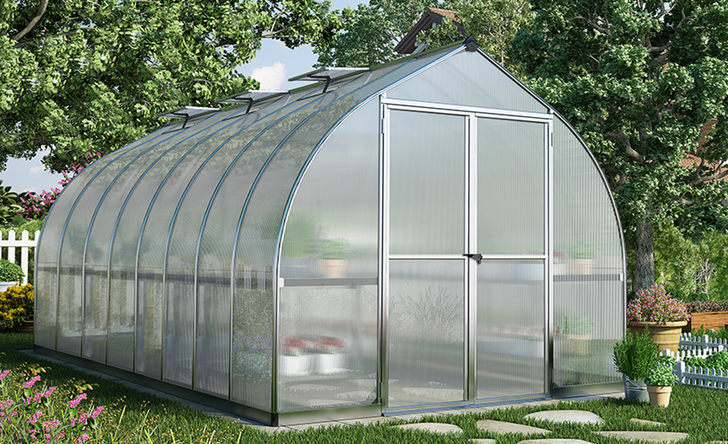 A stone path leads to the front door of a backyard greenhouse covered with polycarbonate panels.