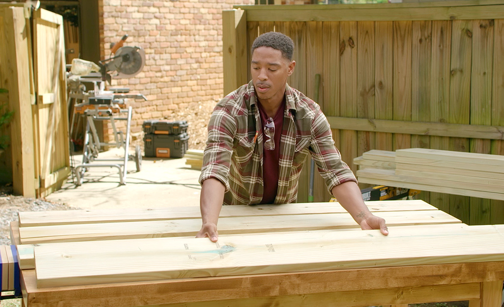 A man assembles the top of a picnic table. A wooden fence and sliding compound miter saw can be seen in the background.