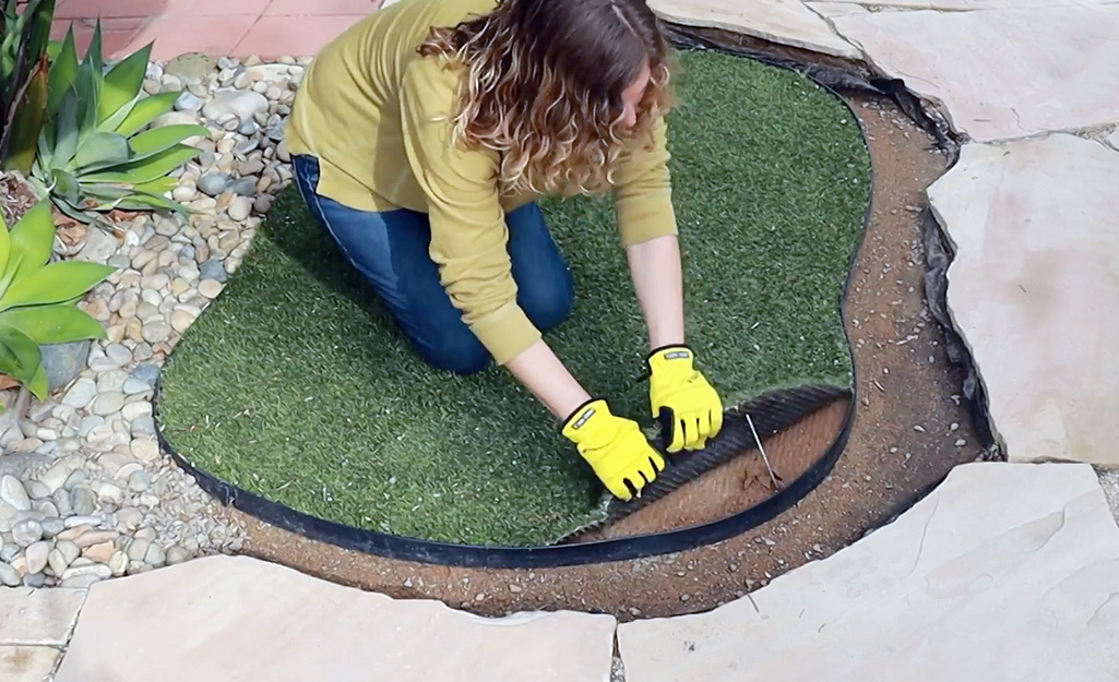 A woman wearing gloves kneels as she pulls up a layer of artificial turf from a small space in a yard.
