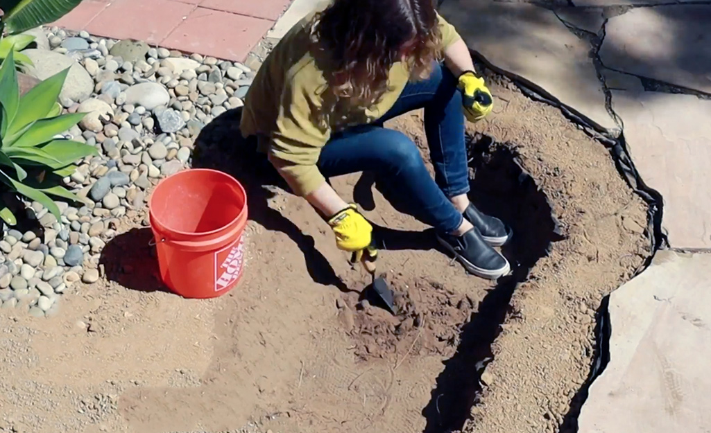 A woman sits on the ground as she uses a trowel to dig out dirt to build a pond.
