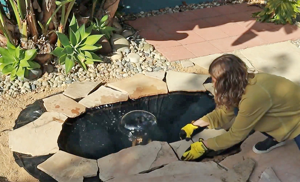 A woman places paver stones around the edge of a small pond to conceal the pond liner.