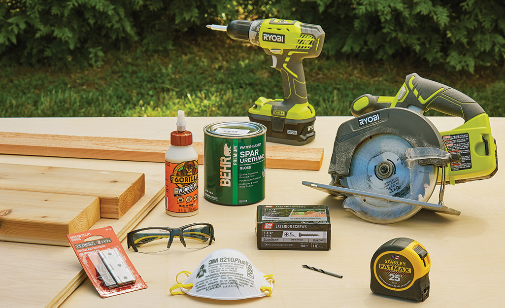 Building supplies including a power drill, screws, saw, tape measure and safety glasses on a table.