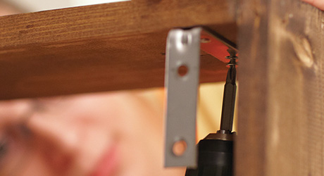 Someone attaching mounting plates to the underside of a wood shelf.