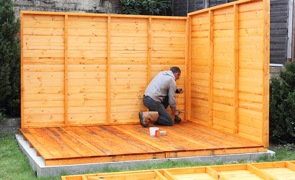 A man installs the back and side walls of a wood shed.