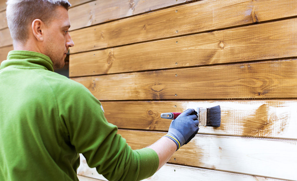 A man staining the side of a wood shed.