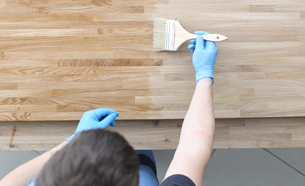 A person uses a broad paint brush to apply stain to a piece of wood.