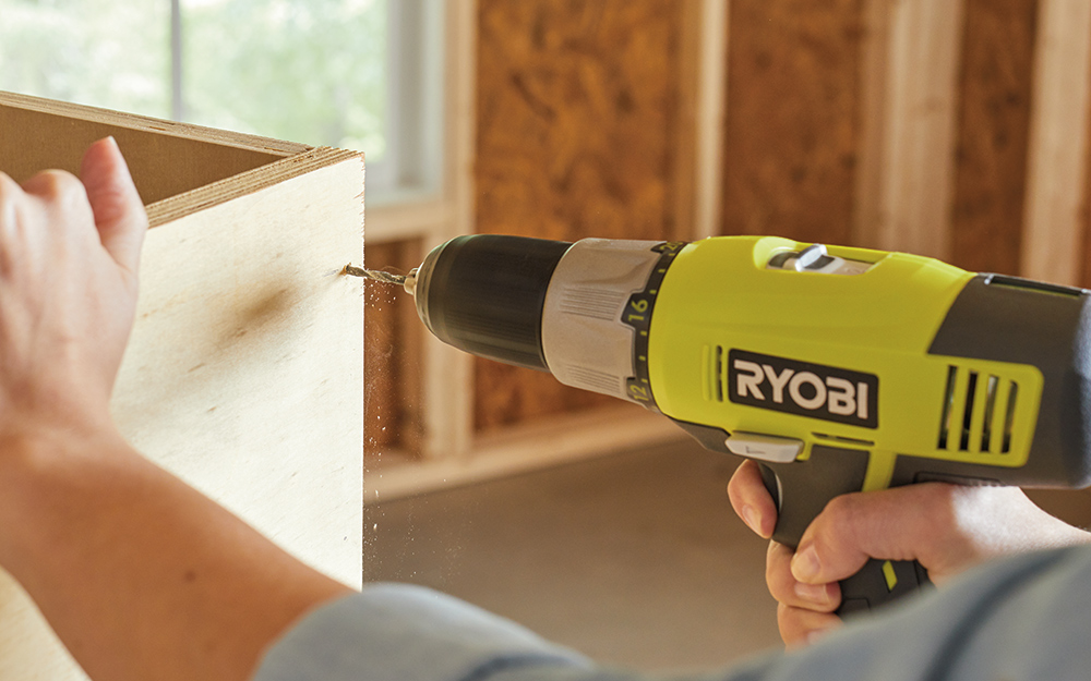 A person using a power drill to pre-drill holes in plywood.