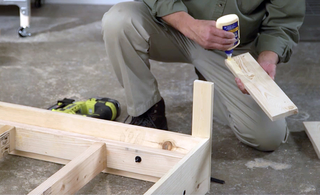 A man applies wood glue to the inner surface of the bed’s legs before nailing them onto the frame