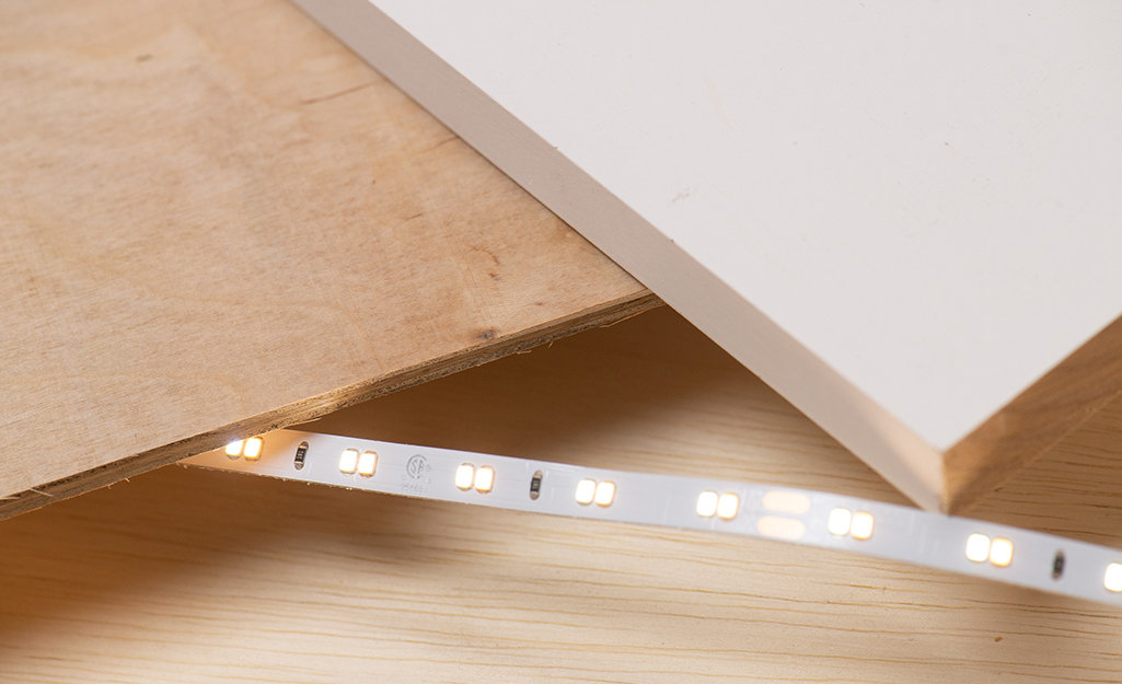 A board, piece of plywood, and section of LED tape strip that are used in the wooden headboard project.