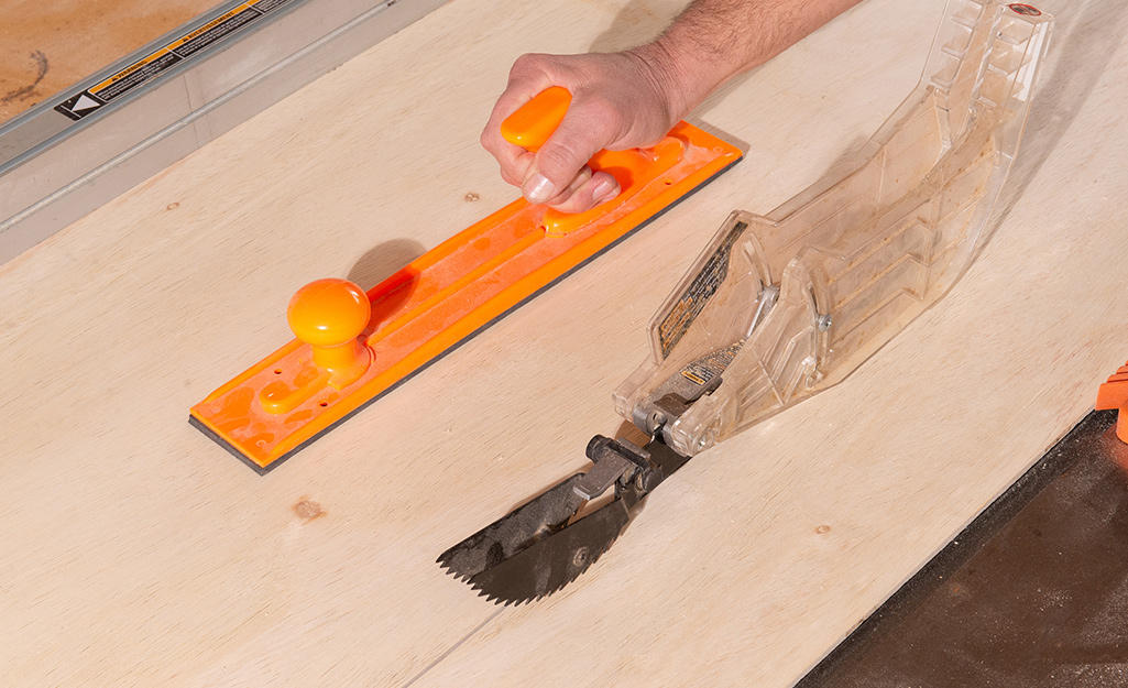 A sheet of plywood being cut on a table saw with the use of a safety push pad. Anti-kickback prawls, riving knife, and blade guard are also visible safety features.