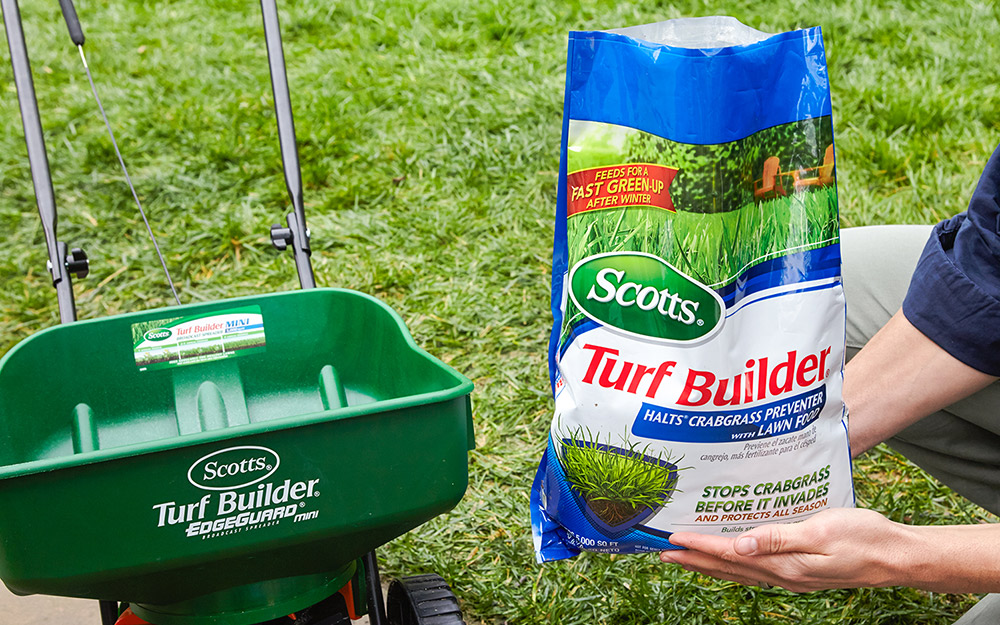 Person holding a bag of fertilizer next to a fertilizer spreader.