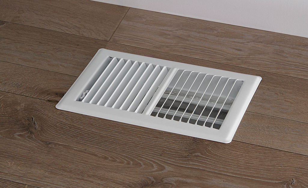 A white vent cover sits on a wooden floor. 