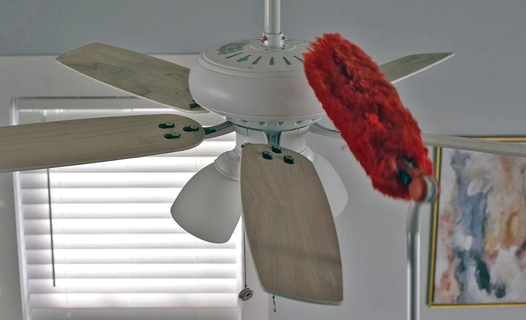 A person dusts a ceiling fan with a duster attached to a pole extension.