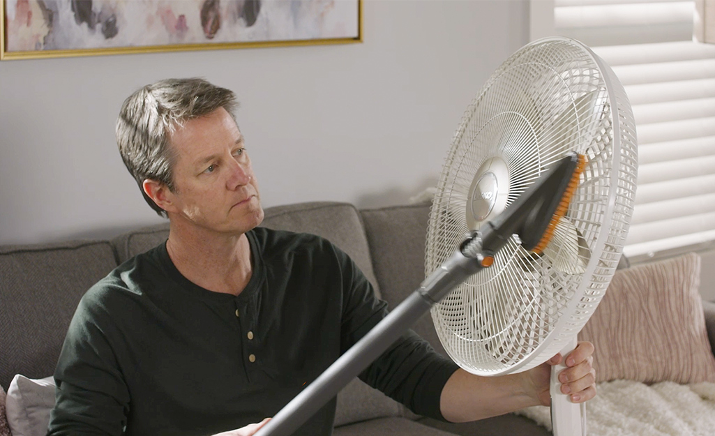 A person vacuums dust and dirt from the grill of an oscillating fan.