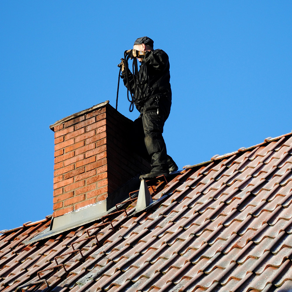 A person stands on a roof uses a rope brush to clean a chimney.