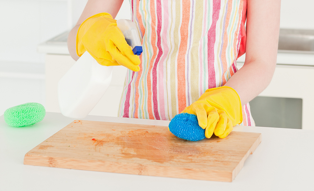 A woman in a kitchen sprays a stained wooden cutting board with cleaner.