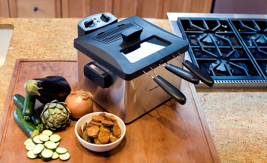 Eggplant, zucchini and other food sits next to a deep fryer on a wooden cutting board in a kitchen.