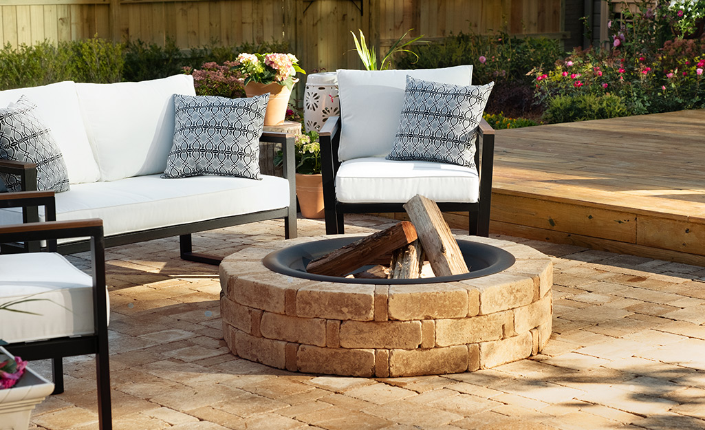 A stone fire pit stands on top of a stone patio and in front of outdoor chairs and a loveseat.