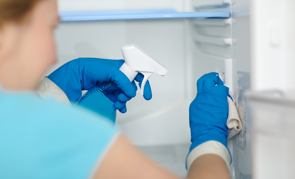 A person wears kitchen gloves to clean a freezer with a cloth and cleaning solution in a spray bottle.
