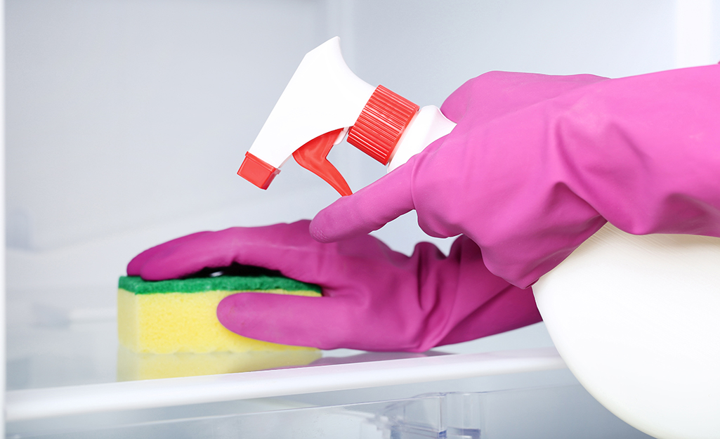 A person wears gloves to clean the inside of a freezer with a sponge and a solution in a spray bottle.