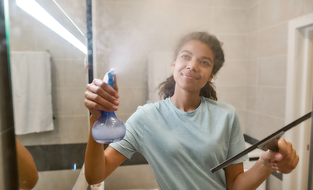 Woman sprays cleaner on to a glass shower door with one hand and holds a squeegee in the other