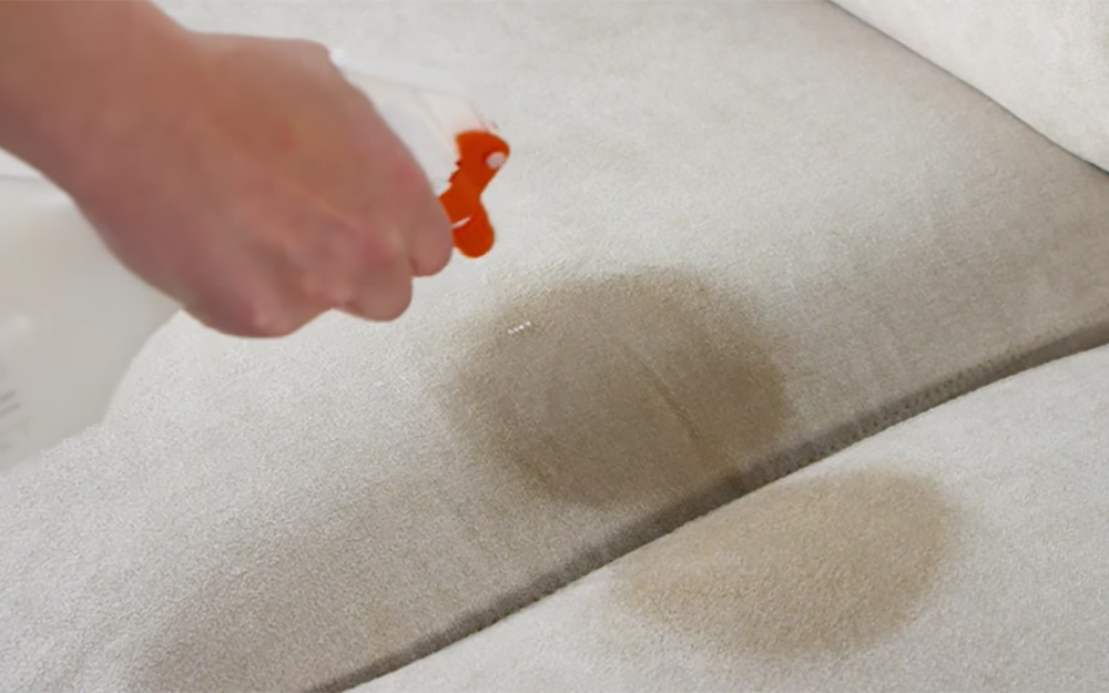 A person sprays a cleaning solution to a stain on a microfiber couch.