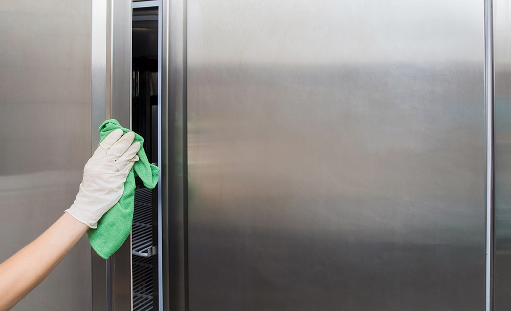 A person wears a glove and uses a cleaning cloth to wipe down the door handle of a refrigerator.