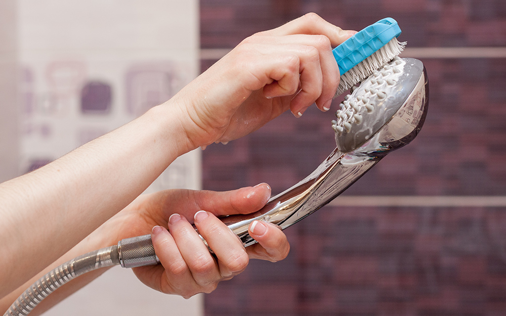 A person cleans a handheld shower head with a scrub brush. 