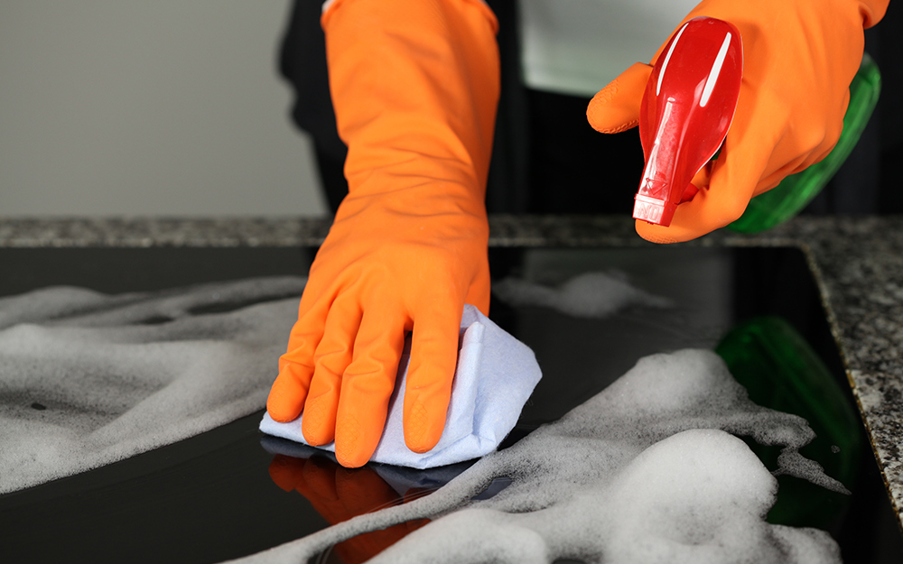 A person wearing orange gloves cleans a ceramic stove top.