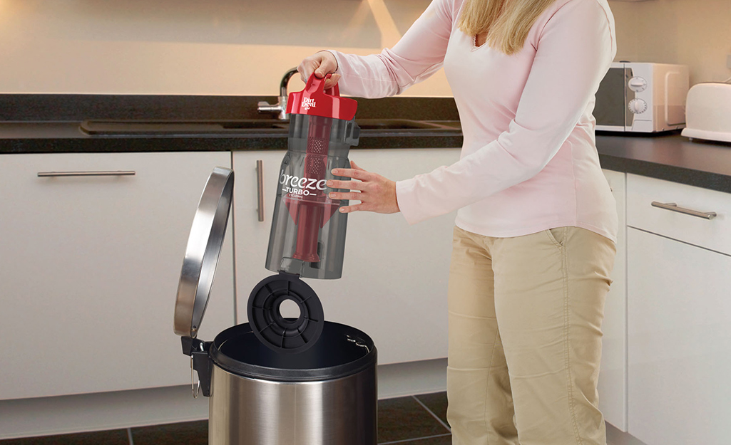 A woman empties a vacuum cleaner canister into a trash can.