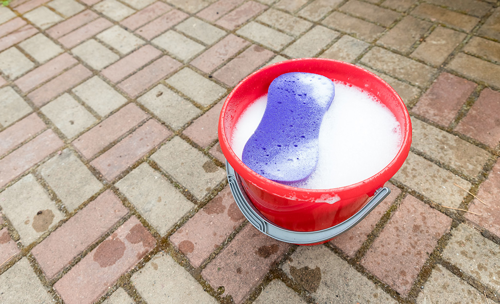 A purple sponge floats in a bucket of cleaning solution on a brick patio.