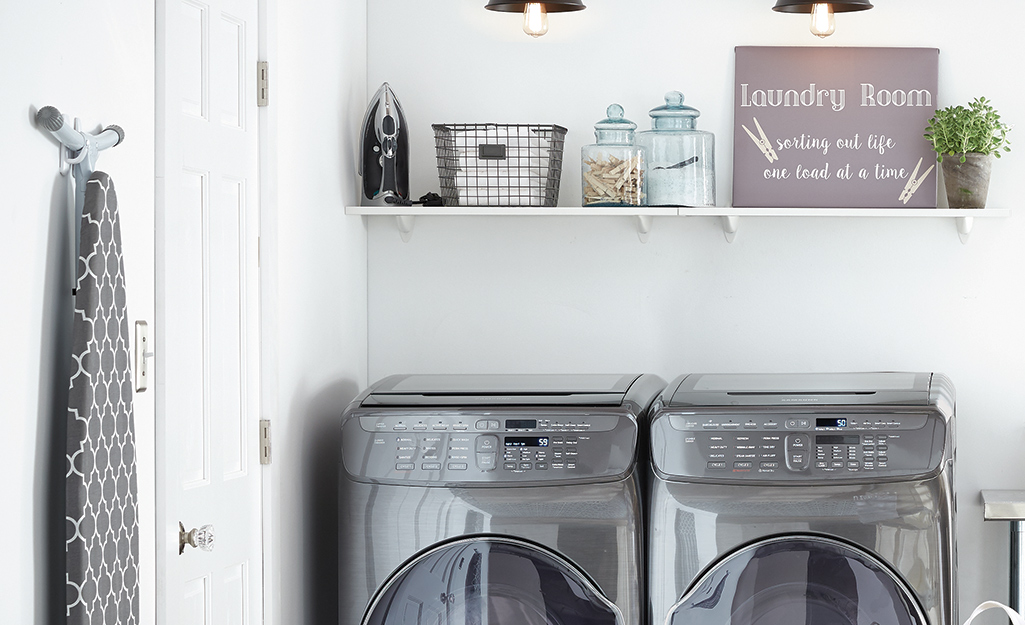 An iron stands upright on a laundry room shelf above a stainless steel washer and dryer.